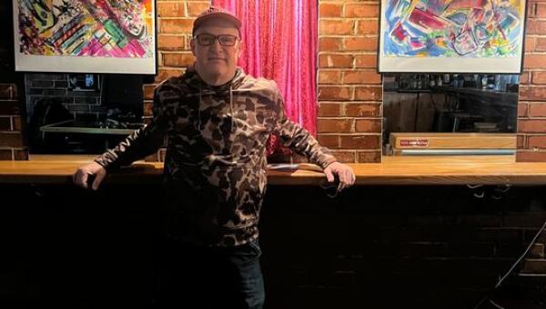 Geo Sangiovanni standing inside Flask Lounge with his artwork behind him.
