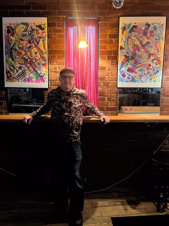 Geo Sangiovanni standing inside Flask Lounge with his artwork behind him.